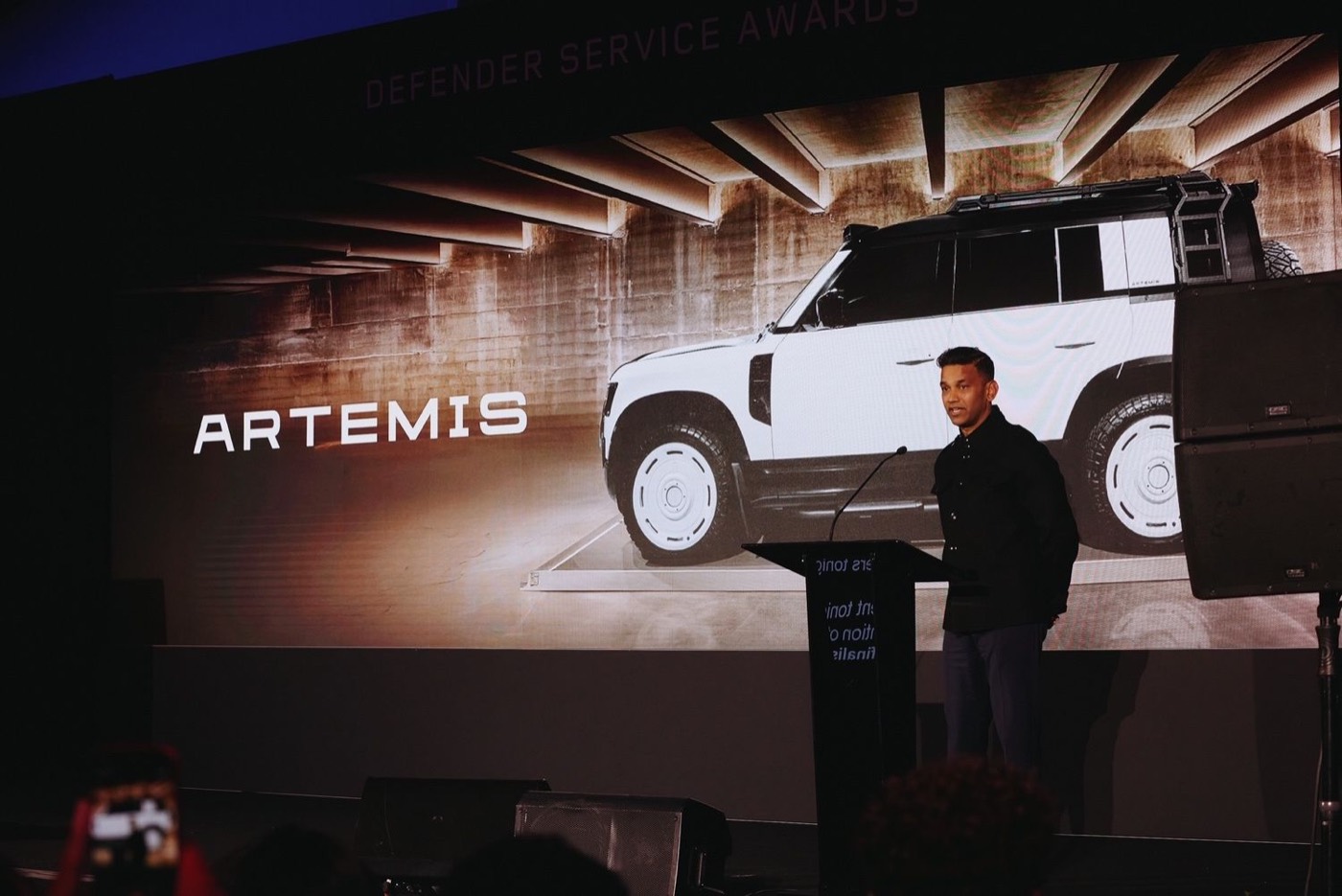 Artemis on stage at the Defender Service Awards, an Artemis-built Land Rover Defender on the screen behind